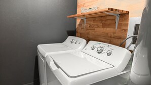 Une buanderie avec deux machines à laver à chargement frontal blanches, une étagère en bois au-dessus et un mur gris.
