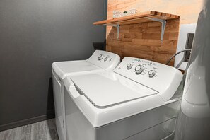 Une buanderie avec deux machines à laver à chargement frontal blanches, une étagère en bois au-dessus et un mur gris.