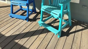 Two Adirondack chairs on a wooden deck in front of a house with large windows.