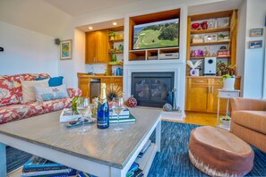 A living room with a sofa, a coffee table with books and a bottle, a fireplace, and a television. - Panoramic OBX 4bed luxury w/ 360° Views, Golf, Beach Club, Pool & Cinema room! (Nags Head)