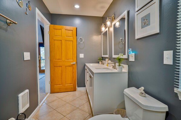A bathroom with a wooden door, a white sink, a mirror, and a toilet. - Panoramic OBX 4bed luxury w/ 360° Views, Golf, Beach Club, Pool & Cinema room! (Nags Head)