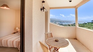 Ein Balkon mit Holztisch und -stühlen, ein Bett mit gestreifter Bettdecke und Blick auf eine Küstenlandschaft.