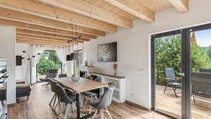 A modern interior with a wooden ceiling, a dining area with a table and chairs, a kitchen area with a stove, and a large sliding glass door leading to an outdoor deck.
