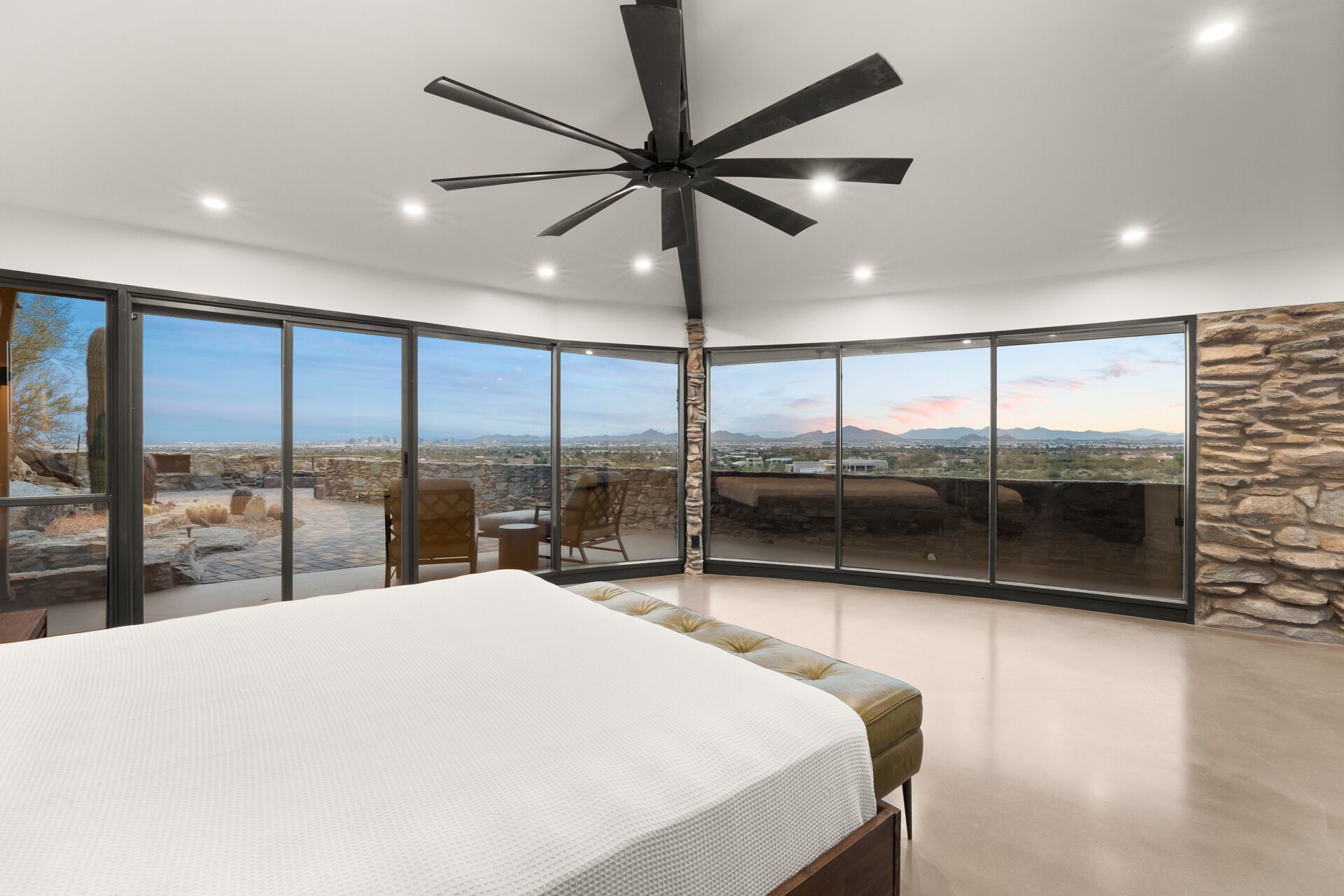 A bedroom with a large bed, a ceiling fan, and a view of a desert landscape through floor-to-ceiling windows.