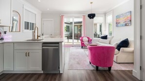 Une cuisine moderne avec des meubles blancs, un évier en acier inoxydable et une chaise rose dans l’espace salon.
