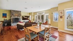 A spacious living room with a stone fireplace, a dining area with wooden furniture, and large windows offering a view of the outdoors. - Cody's Clubhouse: 3 br, wood fireplace, firepit & deck (Waterbury Center)