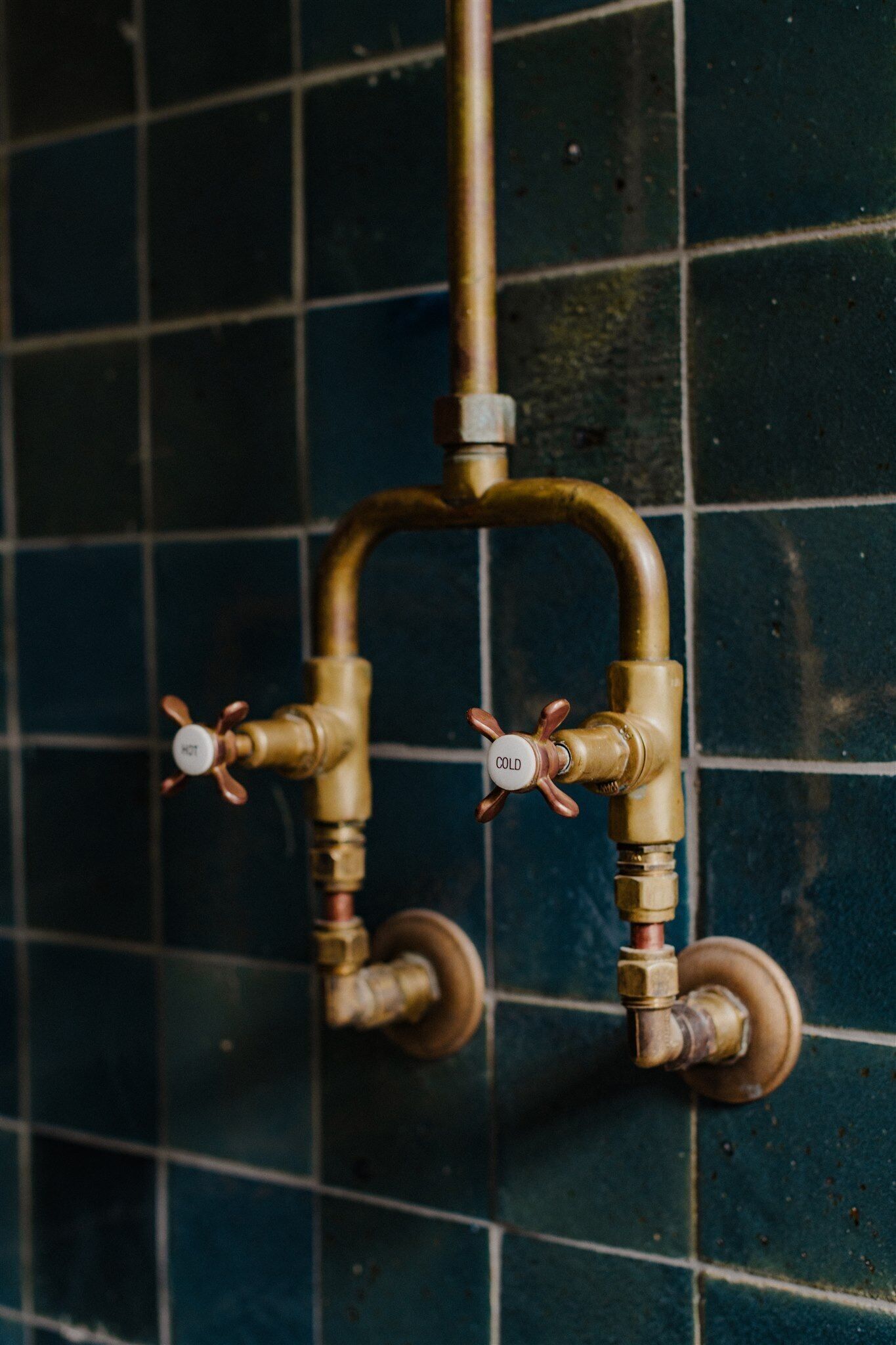 A bathroom with a tiled wall and two brass faucets with temperature control knobs.