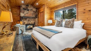 A cozy log cabin bedroom with a stone fireplace, a large bed, and a window overlooking the forest.