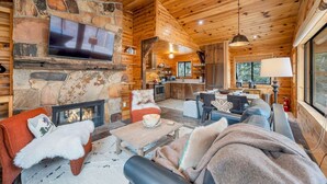 A cozy log cabin interior with a stone fireplace, a TV mounted above it, a dining area with a table and chairs, and a kitchen in the background.