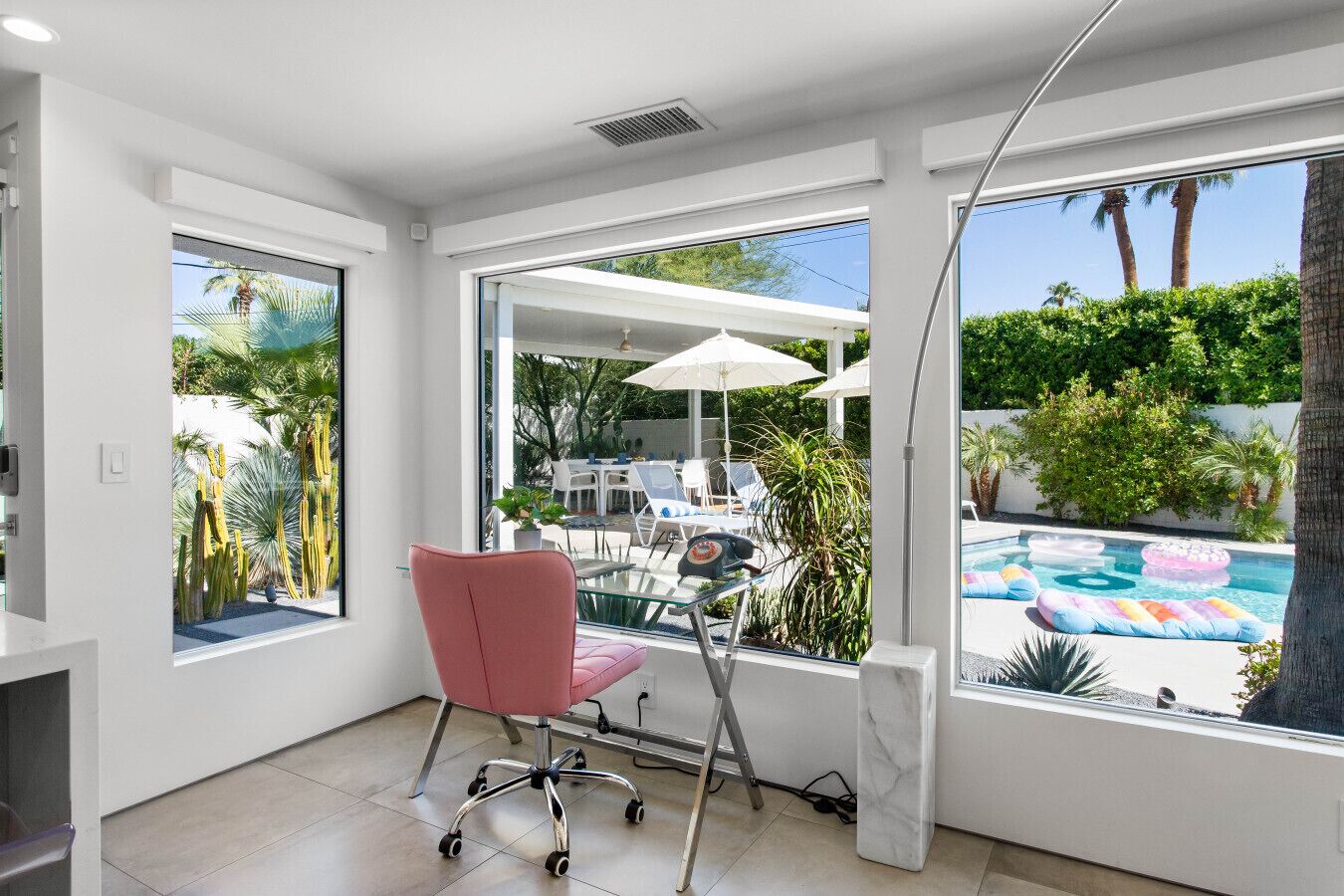 Una habitación moderna con un ventanal grande, una silla de oficina rosa, un escritorio y vistas a un área de piscina exterior con sombrillas y sillones de descanso.