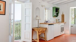 Eine Küche mit weißen Schränken, einem Holztisch, einem grünen Hängeregal und einem Balkon, der durch eine geöffnete Tür zu sehen ist.
