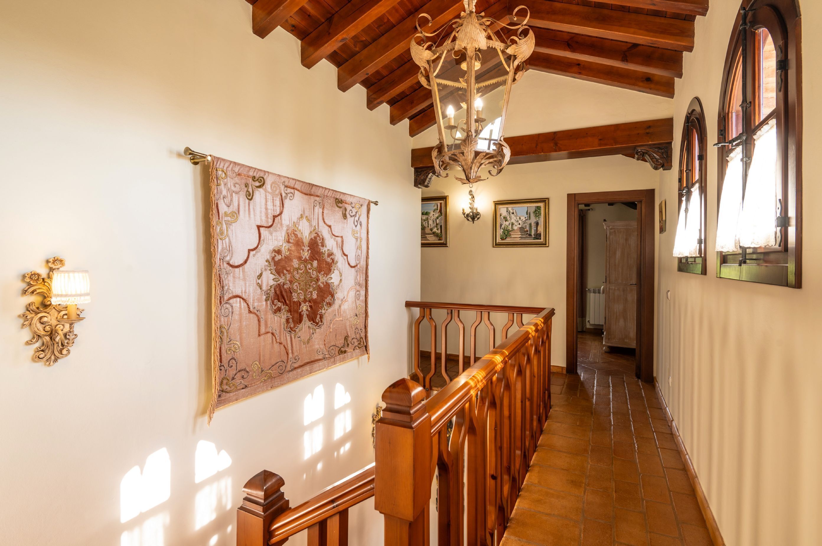 A hallway with a wooden staircase, a chandelier, and a large tapestry on the wall.