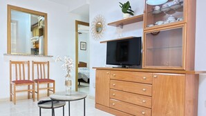 Ein Zimmer mit Fernseher, Holzschrank, zwei Stühlen, einem kleinen Tisch und einem Spiegel.