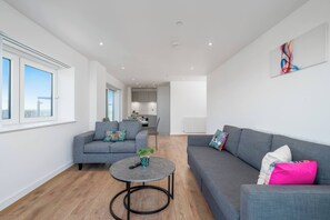 A modern living room with a grey sofa, a round coffee table, and a view of the kitchen in the background. - Skyvillion - River Thames Apt Nr Excel o2 City Ap (London)