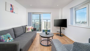 A modern living room with a grey sofa, a round coffee table, a flat-screen TV, and a balcony with a city view. - Skyvillion - River Thames Apt Nr Excel o2 City Ap (London)