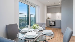 A modern dining area with a glass table, grey chairs, and a view of the city. - Skyvillion - River Thames Apt Nr Excel o2 City Ap (London)