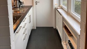 Une cuisine étroite avec des meubles blancs, un évier en acier inoxydable et une fenêtre donnant sur les arbres.