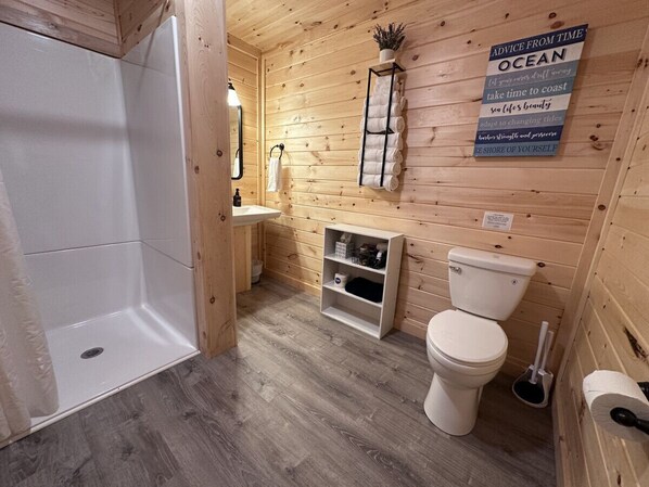 A wooden cabin bathroom with a shower, toilet, sink, and a shelf with toiletries.