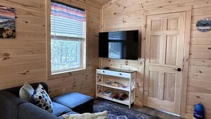 Interior acogedor de una cabaña con techo de madera, un televisor sobre un soporte blanco, un sofá azul con cojines, una ventana con persianas y una puerta.