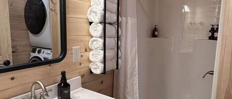 Une salle de bain moderne avec un intérieur en bois, un lavabo blanc doté d’un seul robinet, un porte-serviettes avec des serviettes soigneusement pliées, une douche avec un rideau blanc et un miroir au-dessus du lavabo.
