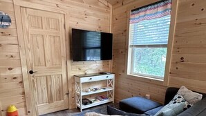 A cozy cabin interior with a wooden interior, a flat-screen TV mounted on a wall, a sofa with cushions, a small white shelf with books, and a window with blinds. - Marshall Lodge. At the heart of Acadia and MDI! (Mount Desert)