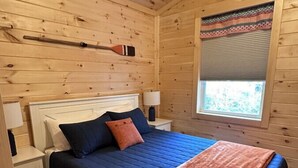 Een slaapkamer in een houten hutje, met een bed, nachtkastjes, een raam en een roeiblad aan de wand.