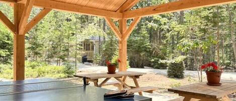A covered outdoor area with a ping pong table, picnic tables, and a wooden structure.