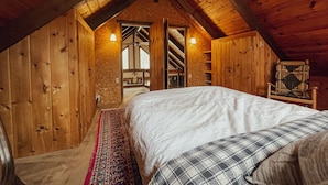 A cozy attic bedroom with a wooden sloped ceiling, a bed with a plaid blanket, a red and blue patterned rug, and a wooden chair. - Cozy chalet near lakes and mountains. Pet friendly! (Lovell)