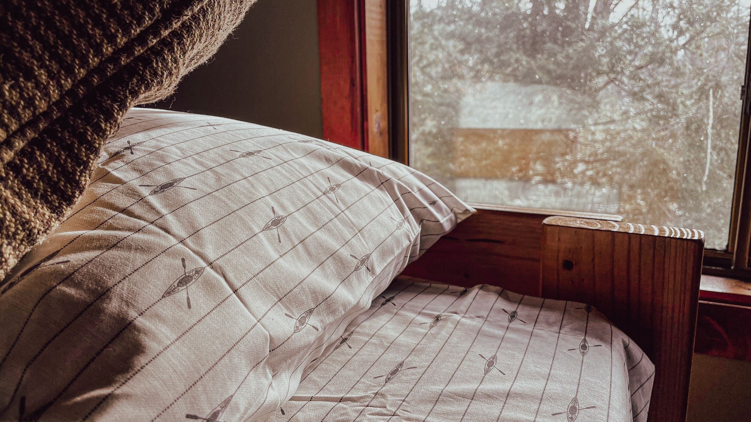 Un lit avec un oreiller à rayures et une tête de lit en bois, placé près d’une fenêtre donnant sur des arbres.