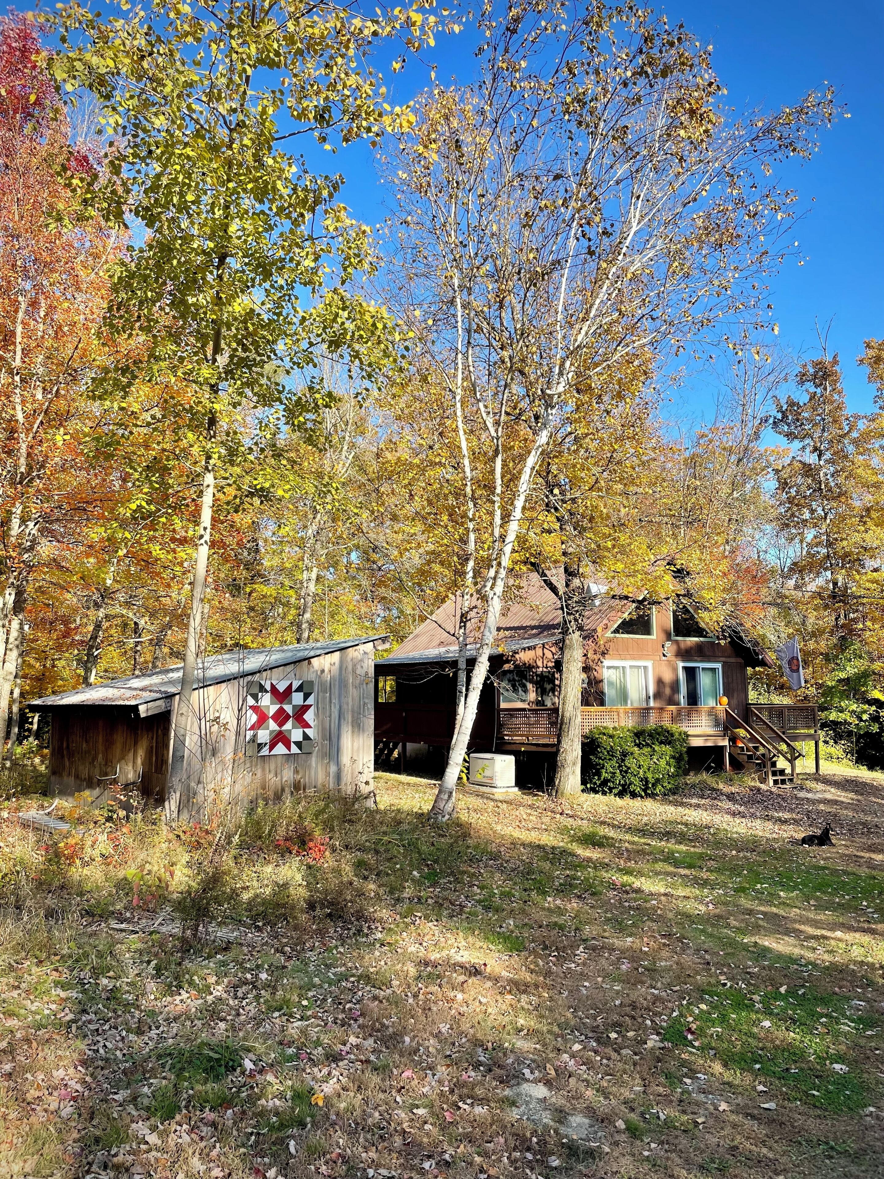 A rustic cabin with a wooden deck and a covered porch, surrounded by autumn foliage.