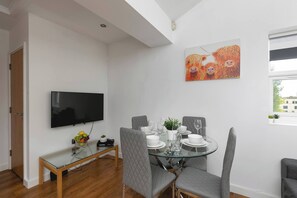 A dining area with a glass table, grey upholstered chairs, a flat-screen TV mounted on the wall, and a painting of dogs. - Skyvillion - Central Ldn 3bed Flat Ladbroke Grove (London)