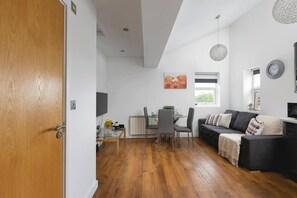 A modern living room with a black sofa, a glass coffee table, and a dining area with a table and chairs. - Skyvillion - Central Ldn 3bed Flat Ladbroke Grove (London)