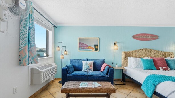 Une pièce avec un canapé bleu, un lit doté d’une tête de lit en osier, une petite table et un ventilateur fixé au mur.