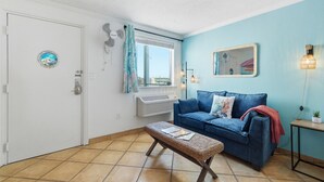 Une pièce avec un canapé bleu, un banc en osier, une porte blanche, une fenêtre avec des rideaux et un ventilateur fixé au mur.