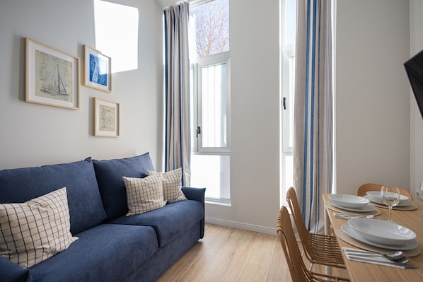 A room with a blue sofa, a dining table set with plates and a glass, and framed artwork on the wall. - Pirate Duplex IV | 4 Guests | AC WiFi | 15 min Oceanografic (Valencia)