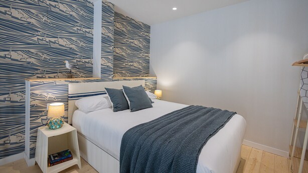 A neatly arranged bedroom with a patterned wall, a bedside table with a lamp, and a bed with pillows and a blanket.