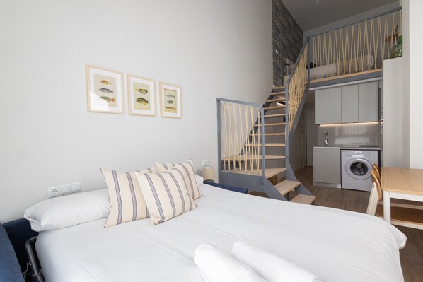 Un dormitorio con cama, almohadas, una escalera, lavadora y cuadros en la pared.
