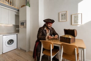 Une personne est assise à une table devant une caisse remplie de pièces de monnaie, dans une pièce où se trouvent un lave-linge, un four à micro-ondes et des dessins encadrés accrochés au mur.