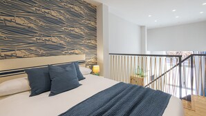 Una habitación de hotel moderna con una cama grande, un cabecero estampado, un balcón con barandilla y una mesita de noche con lámpara.