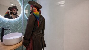 Una persona disfrazada con un loro en un baño moderno con un espejo redondo, un lavabo y una ducha.