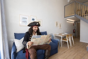 Une personne est assise sur un canapé bleu dans une pièce où se trouvent une table à manger, des chaises et un escalier.