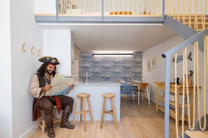 Une personne vêtue d’un costume de pirate est en train de lire une carte dans une pièce dotée d’un plafond haut, de tabourets en bois, d’un bar et d’un escalier en corde.