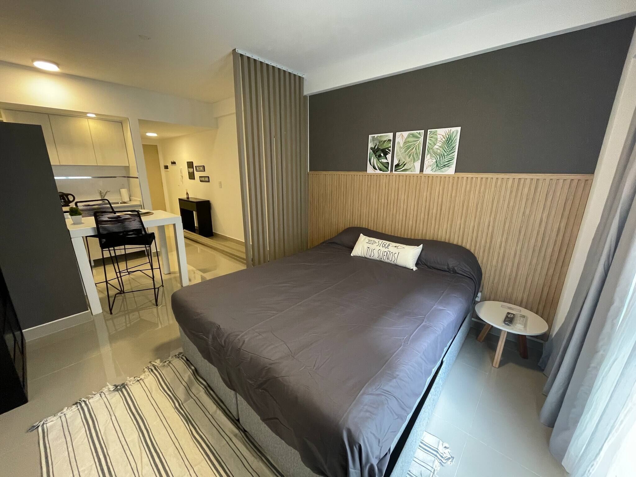A modern hotel room with a large bed, a small desk, and a kitchenette.
