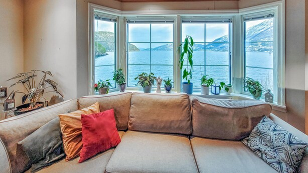 Un cuarto acogedor con un ventanal grande con vista a un lago, un rincón de descanso con cojines y varias plantas en macetas.