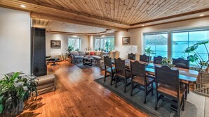 Amplia sala de estar con piso de madera, una mesa de comedor con sillas y ventanas grandes con vista al agua.