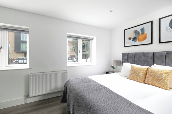 A modern bedroom with a bed, two windows, a radiator, and artwork on the wall.