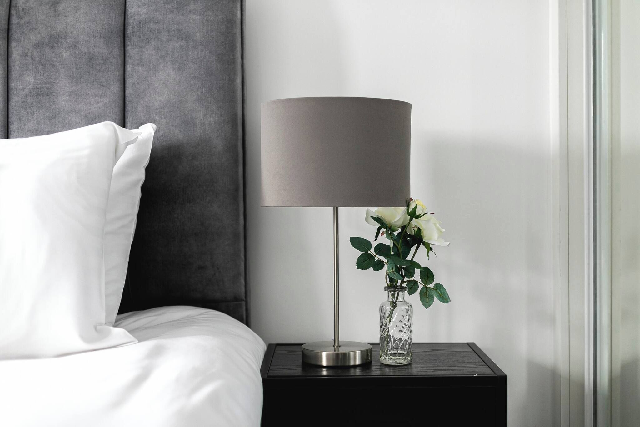 A neatly made bed with white linens, a grey headboard, a modern lamp with a cylindrical shade, and a vase with white flowers on a bedside table.