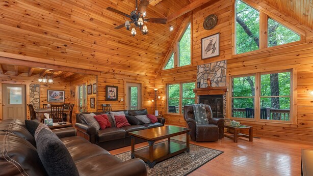 Interior espacioso de una cabaña de madera con una amplia sala de estar, una chimenea y múltiples ventanas que ofrecen vistas al bosque circundante.
