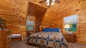 Un dormitorio acogedor con techo de madera, cama con colcha escocesa, mesita de noche con lámpara y ventana con persianas.