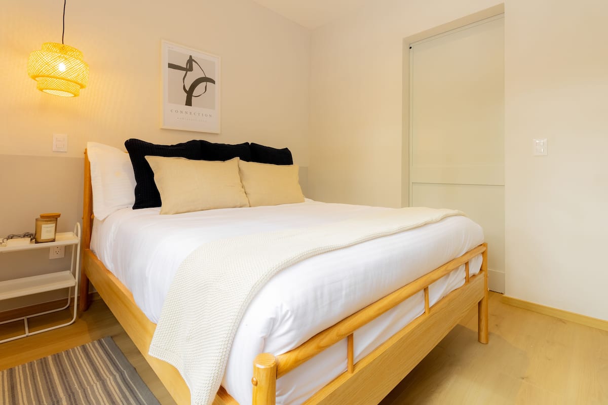 Un letto rifatto con lenzuola bianche e una testiera in legno, un comodino con una lampada e un quadro incorniciato appeso al muro.